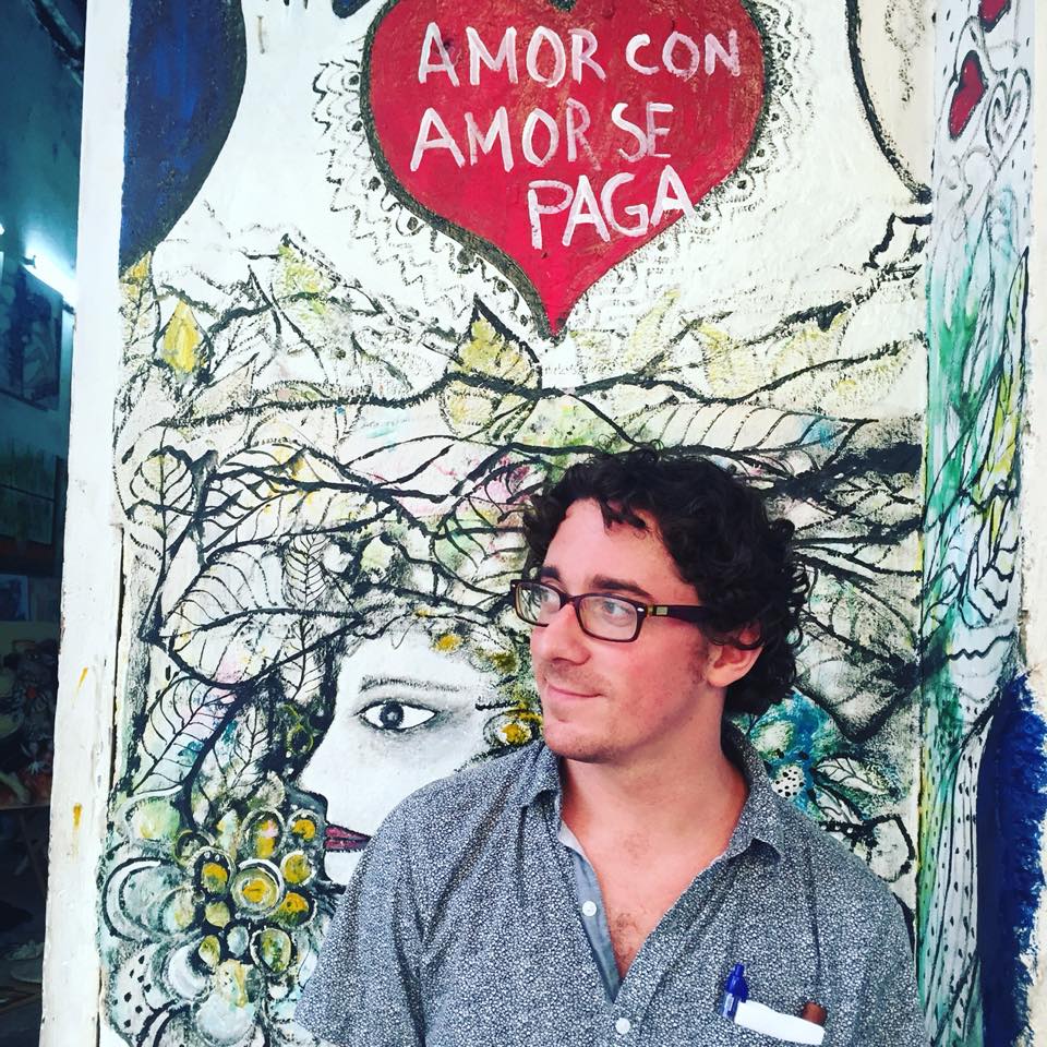 A portrait of an adult man with dark, curly hair and glasses. He is wearing a gray shirt and standing in front of a mural.